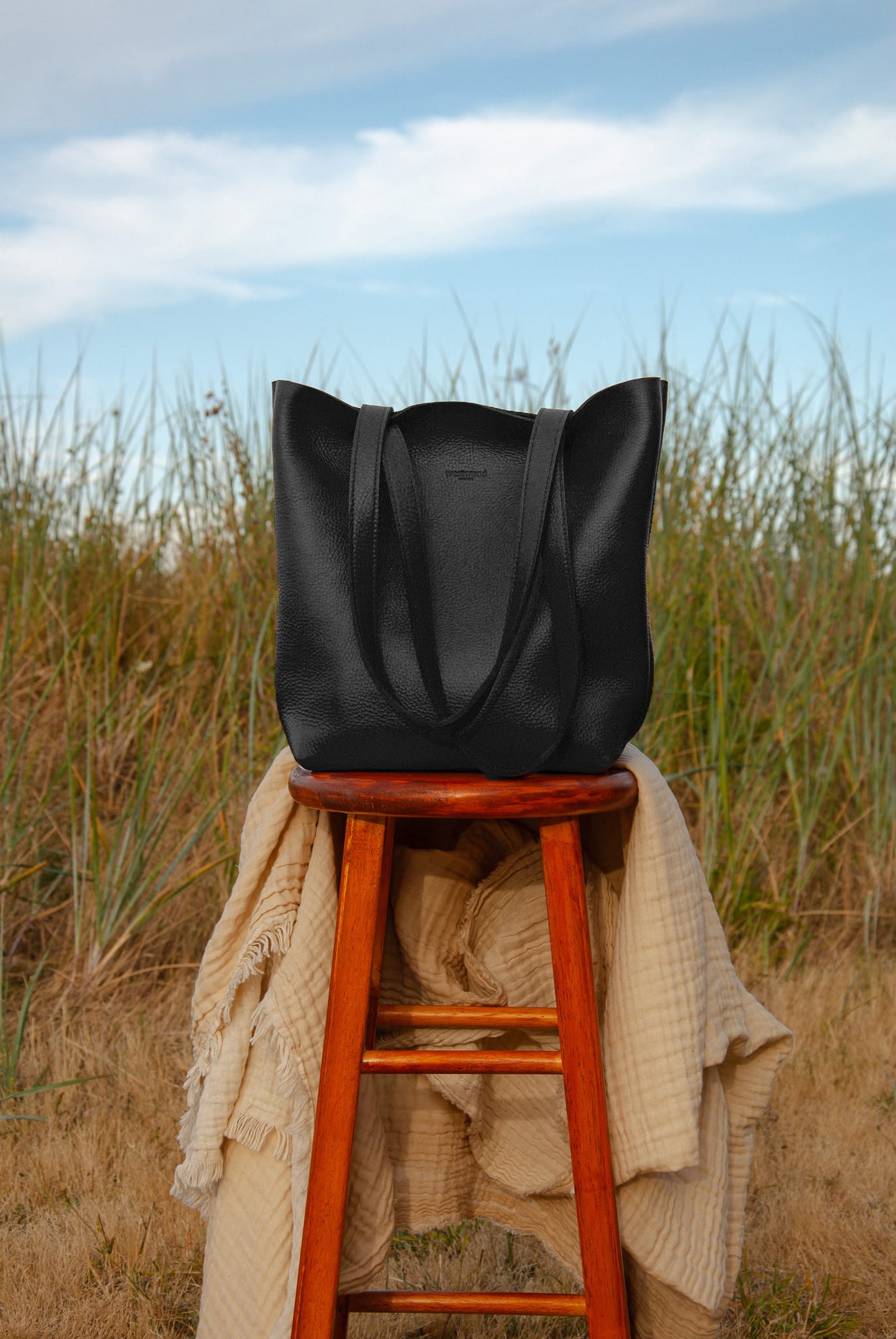 Black tote bag on a wooden stool with grass and blue sky in the background