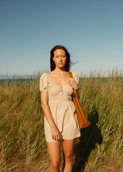 Woman in a beige dress holding a brown bag in a grassy field with a clear blue sky.