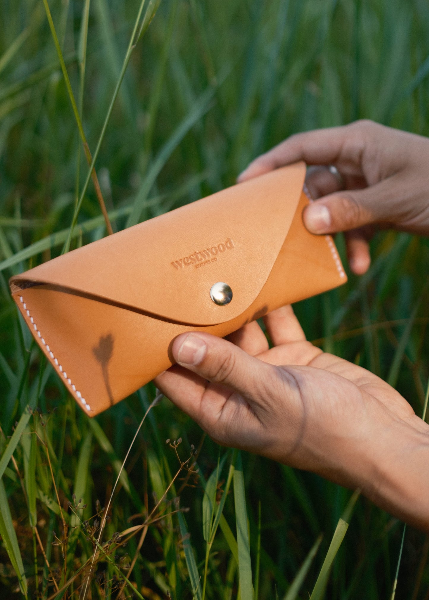 Person holding a brown leather glasses case with 'Westwood' branding in a grassy outdoor setting