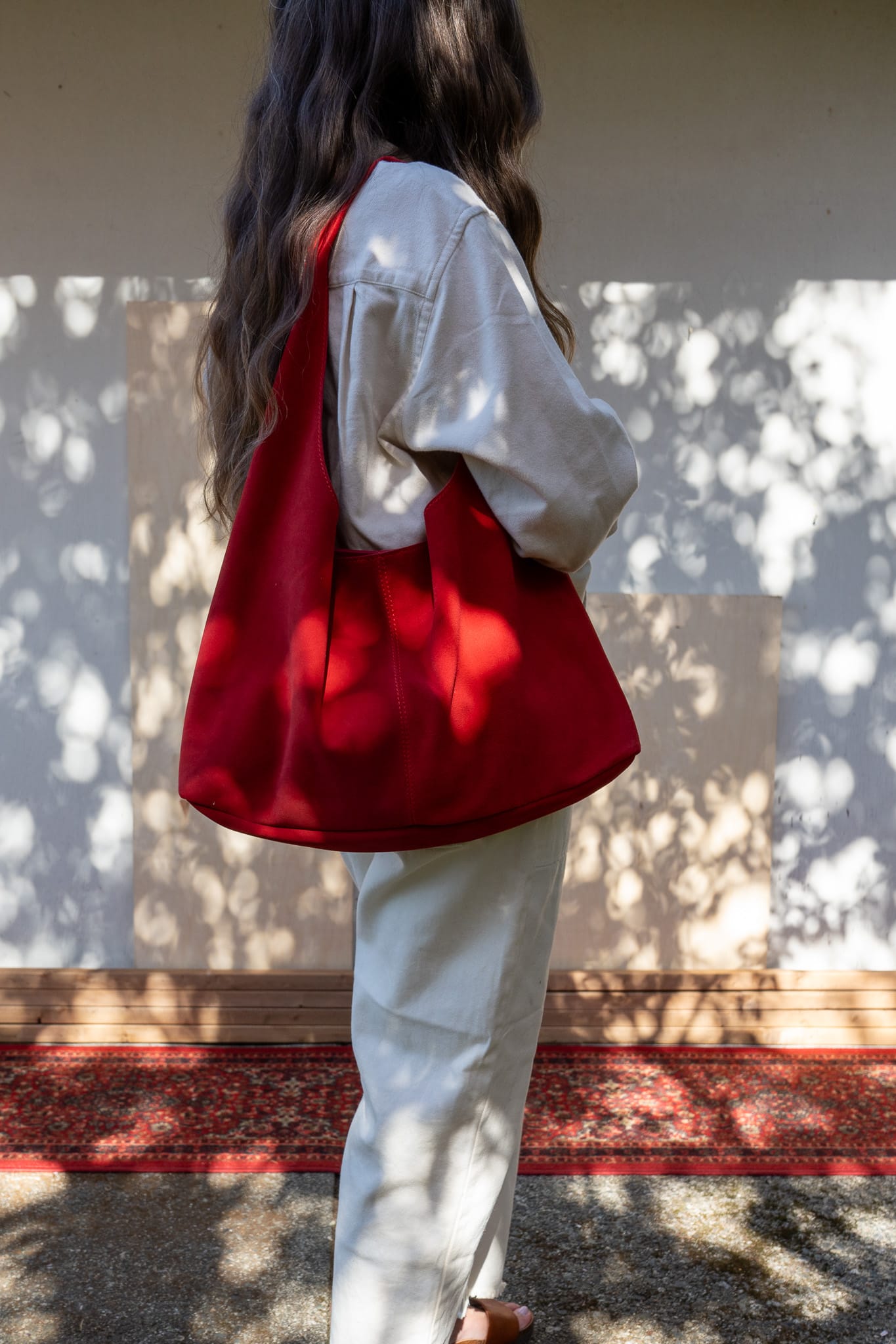 A person viewed from behind carrying a red slouchy tote bag over their shoulder, with a white shirt and light-colored pants.