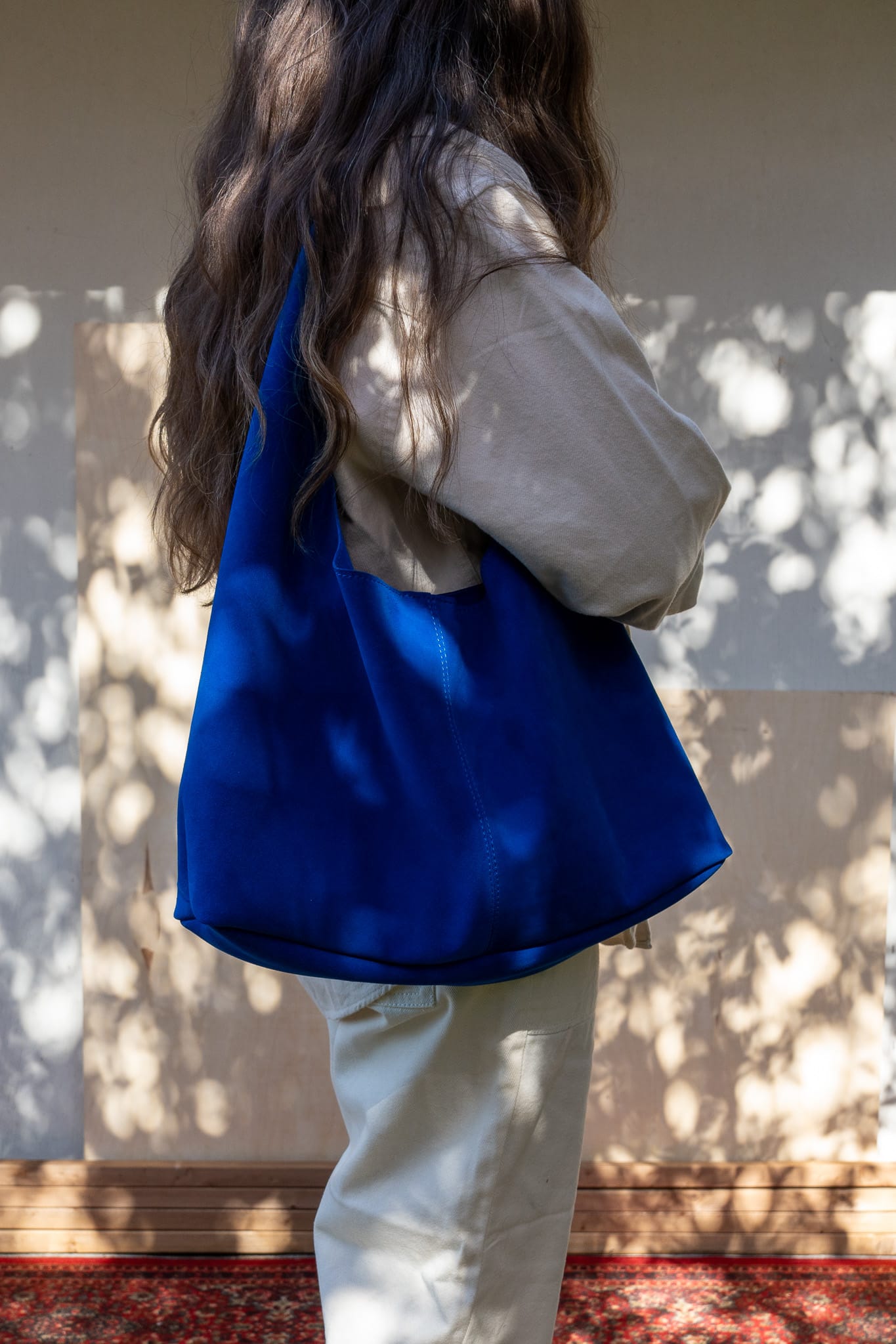 A person viewed from behind carrying a blue slouchy tote bag over their shoulder, with a white shirt and light-colored pants.