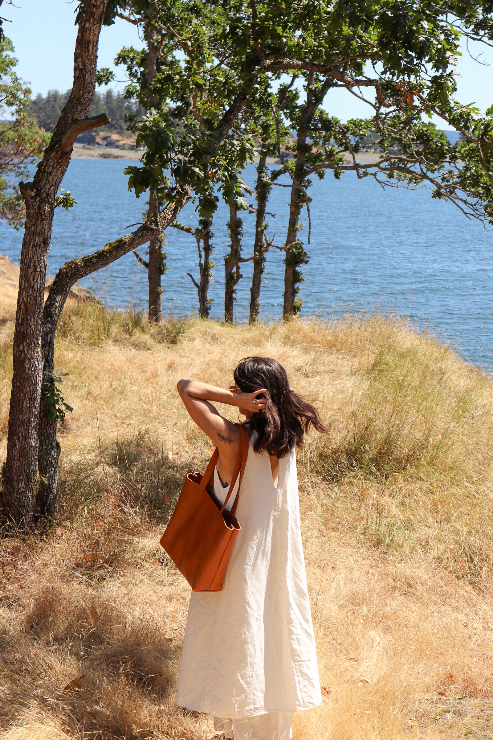 a woman is seen from behind in a long white dress with a brown tote bag outdoors by the ocean