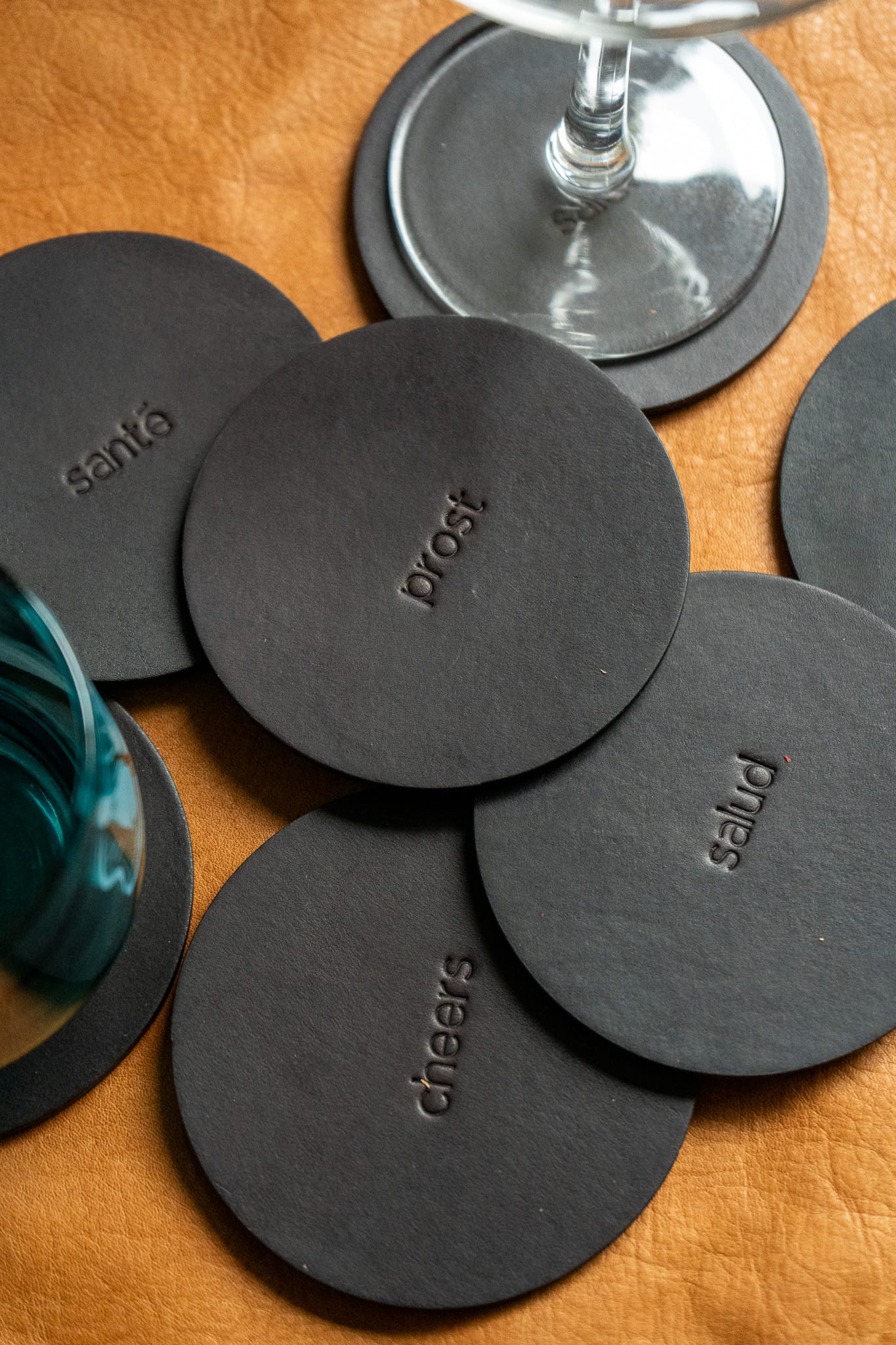 A set of round, black leather coasters  with engraving with a glass in the background.
