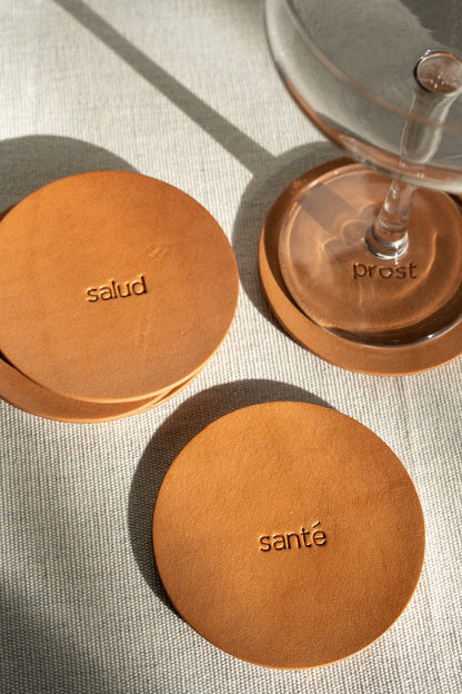 A set of round, brown leather coasters with engraving displayed on a cream surface with a glass in the background.