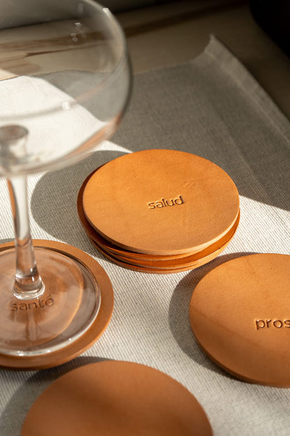 A set of round, brown leather coasters with engraving displayed on a cream surface with a glass in the background.