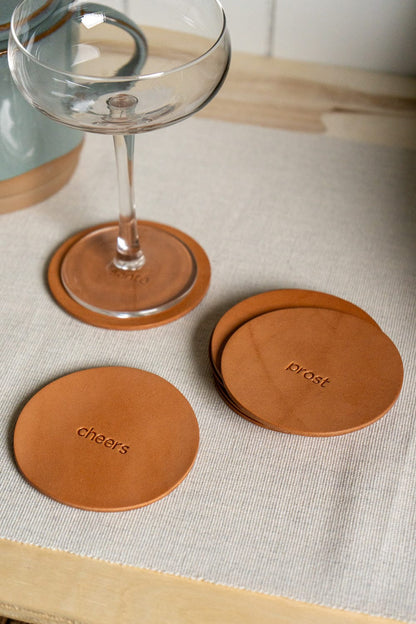 A set of round, brown leather coasters with engraving displayed on a cream surface with a glass in the background.