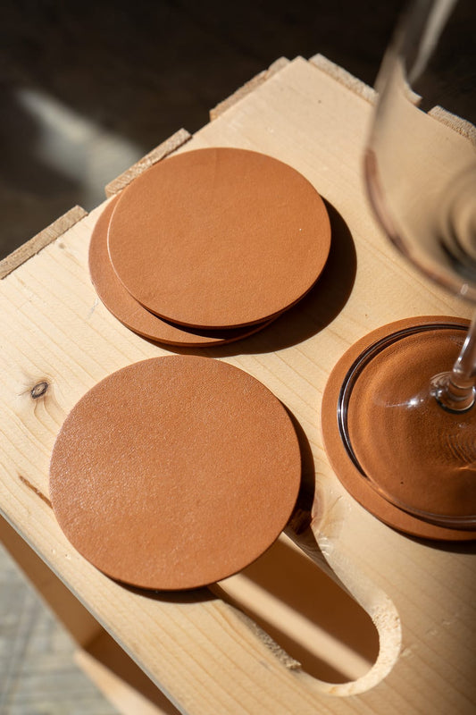 A set of round, brown leather coasters with displayed on a wooden surface with a glass in the background.