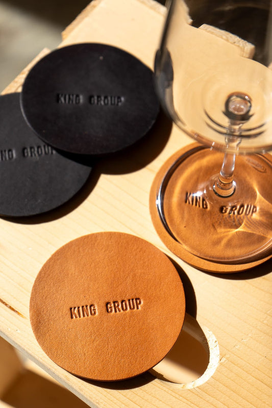 A set of round, brown leather coasters with the engraved text 'KING GROUP' displayed on a wooden surface with a glass in the background.