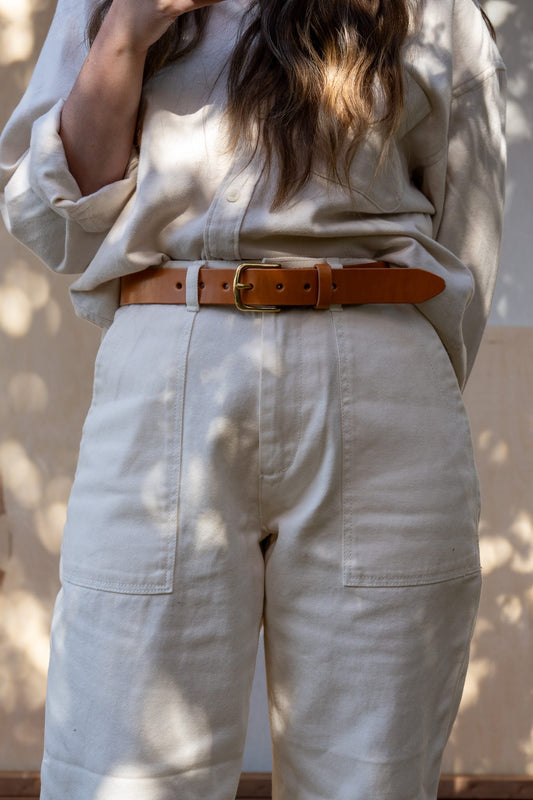 A person wearing a beige belt with a brown buckle and matching leather keeper, standing in a shadowed area with foliage in the background.
