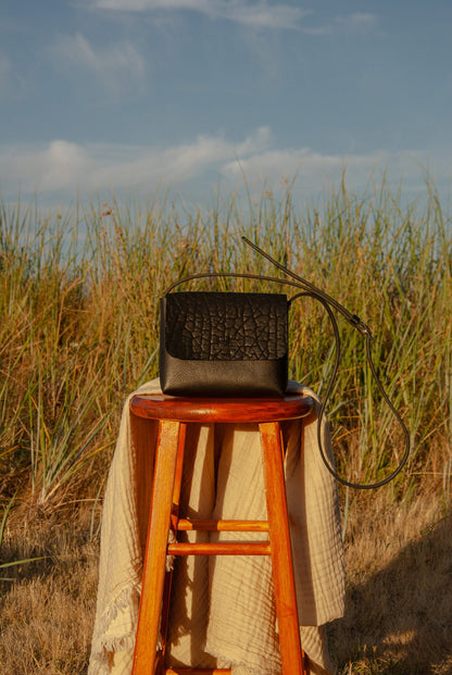 Black handbag on a wooden stool with tall grass and blue sky in the background