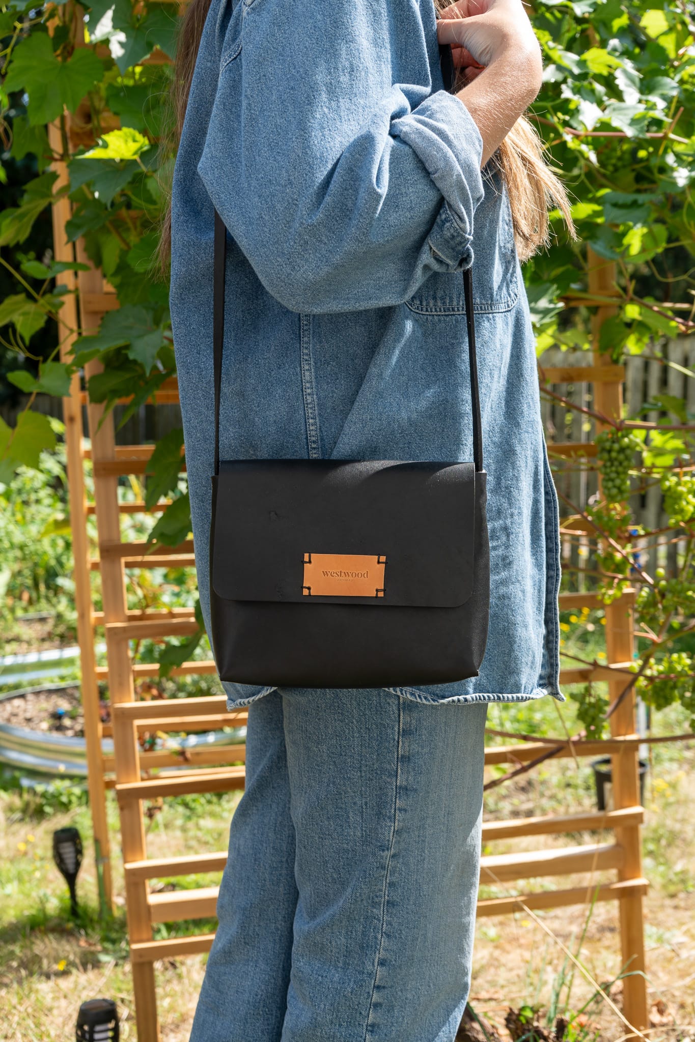 a person wearing a denim shirt and pants stands in front of foliage wearing a black leather purse on their shoulder.