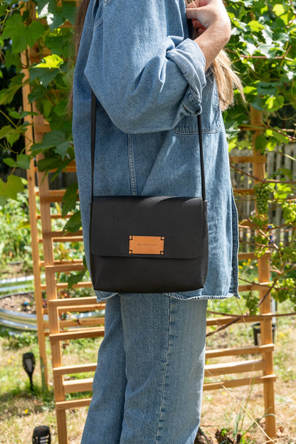 a person wearing a denim shirt and pants stands in front of foliage wearing a black leather purse on their shoulder.