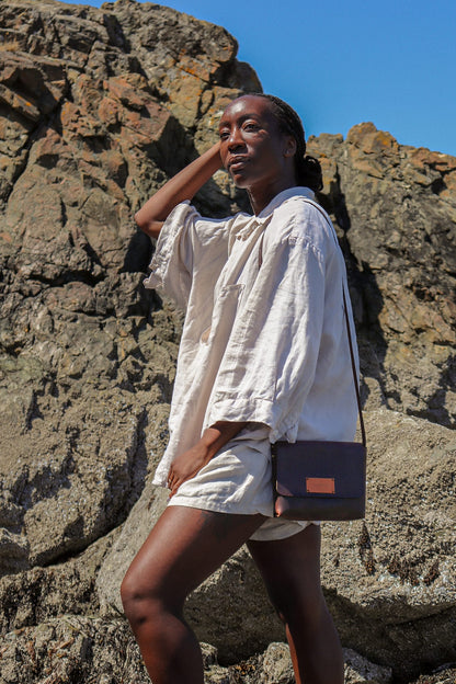 a person wearing a beige shirt stands in front of a rock wall wearing a brown leather purse on their shoulder.