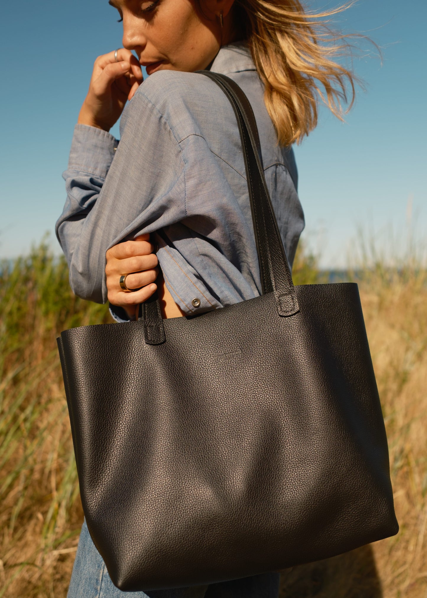 Person holding a black tote bag in an outdoor setting with grass and blue sky.