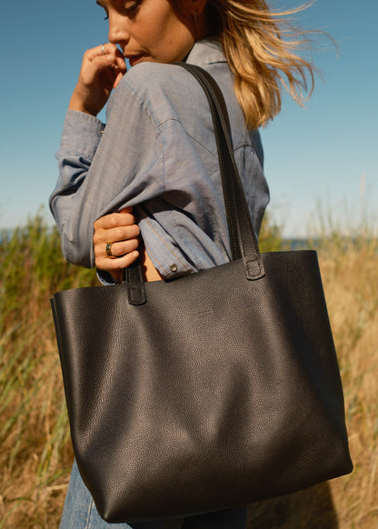 Person holding a black tote bag in an outdoor setting with grass and blue sky.