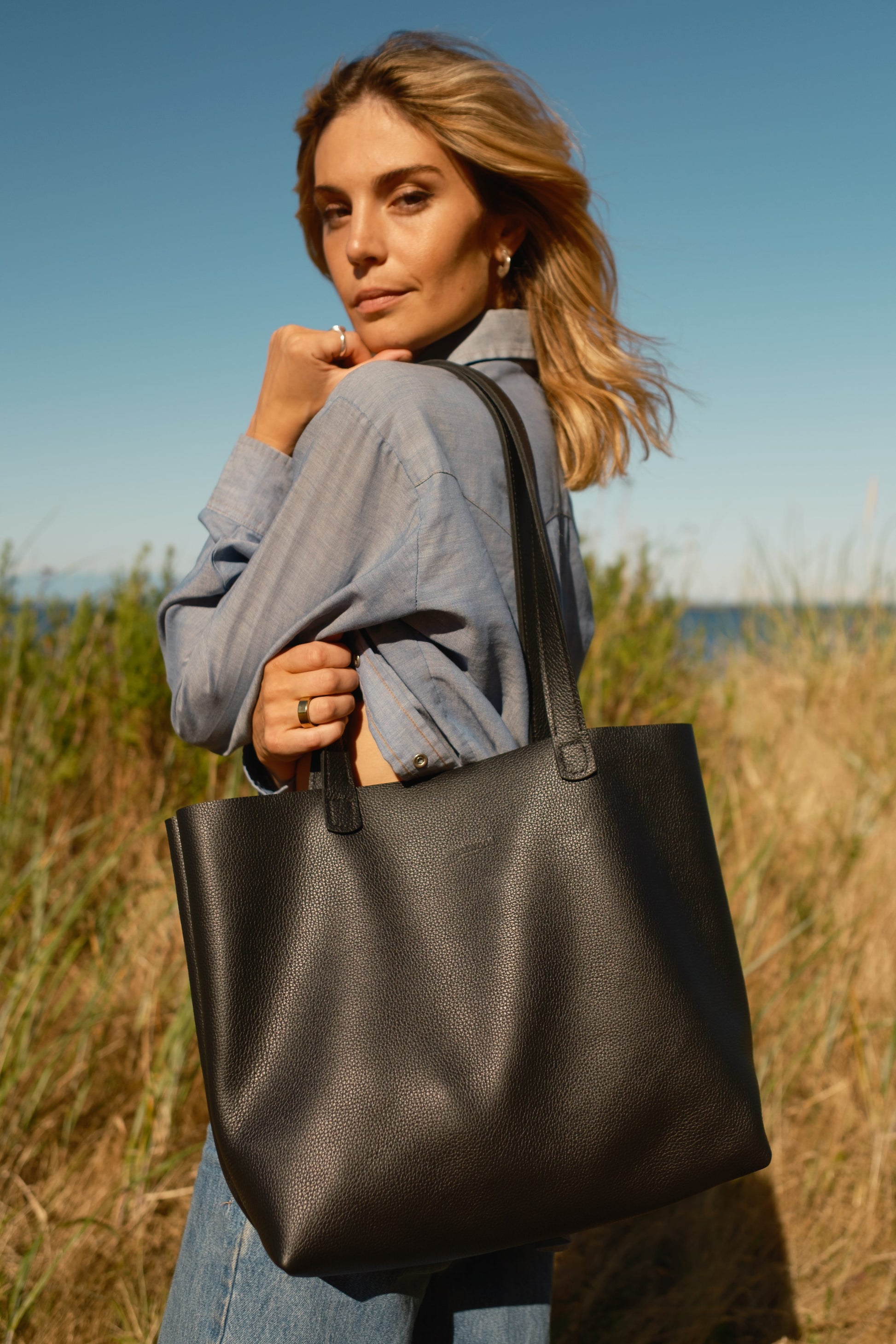 Woman holding a black tote bag in a field with a clear blue sky.