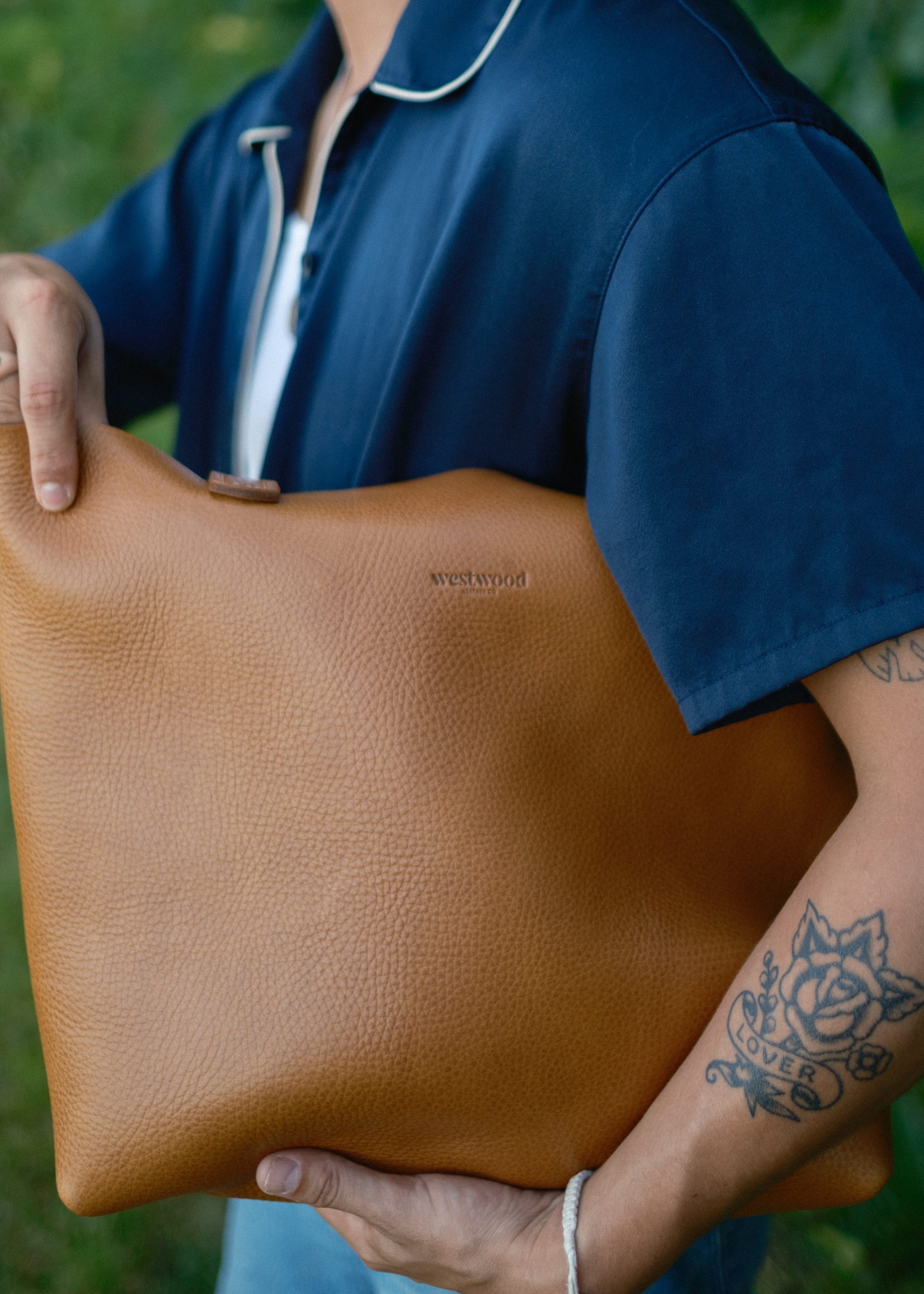 Person holding a brown leather clutch with 'Westwood' branding against a blurred green background