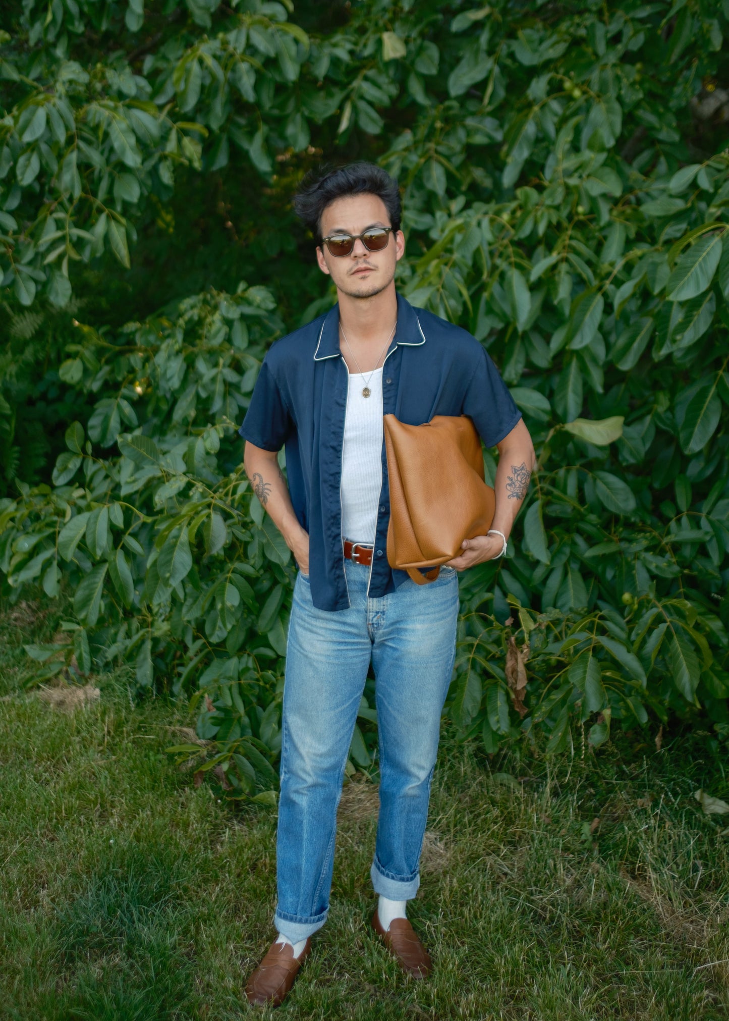 Man holding a brown leather bag in front of green foliage