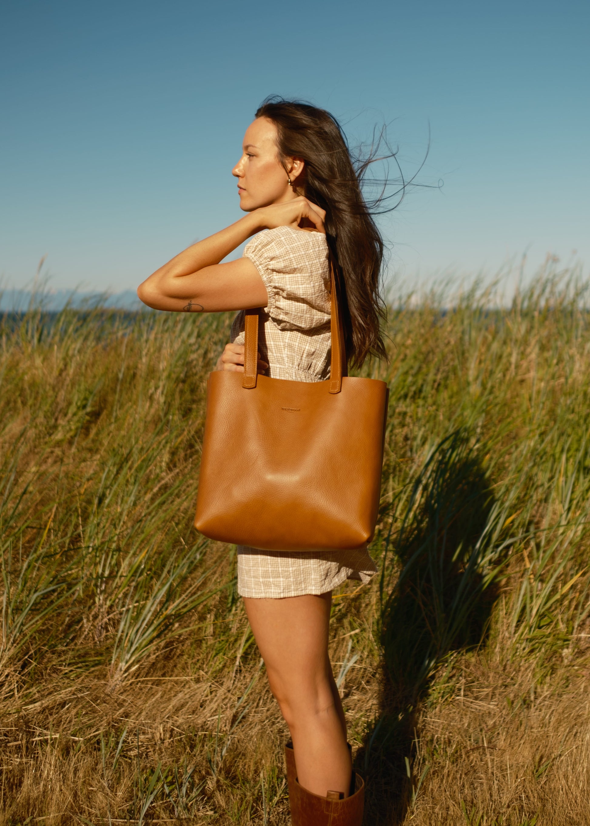 Woman holding a brown leather tote bag in a grassy field