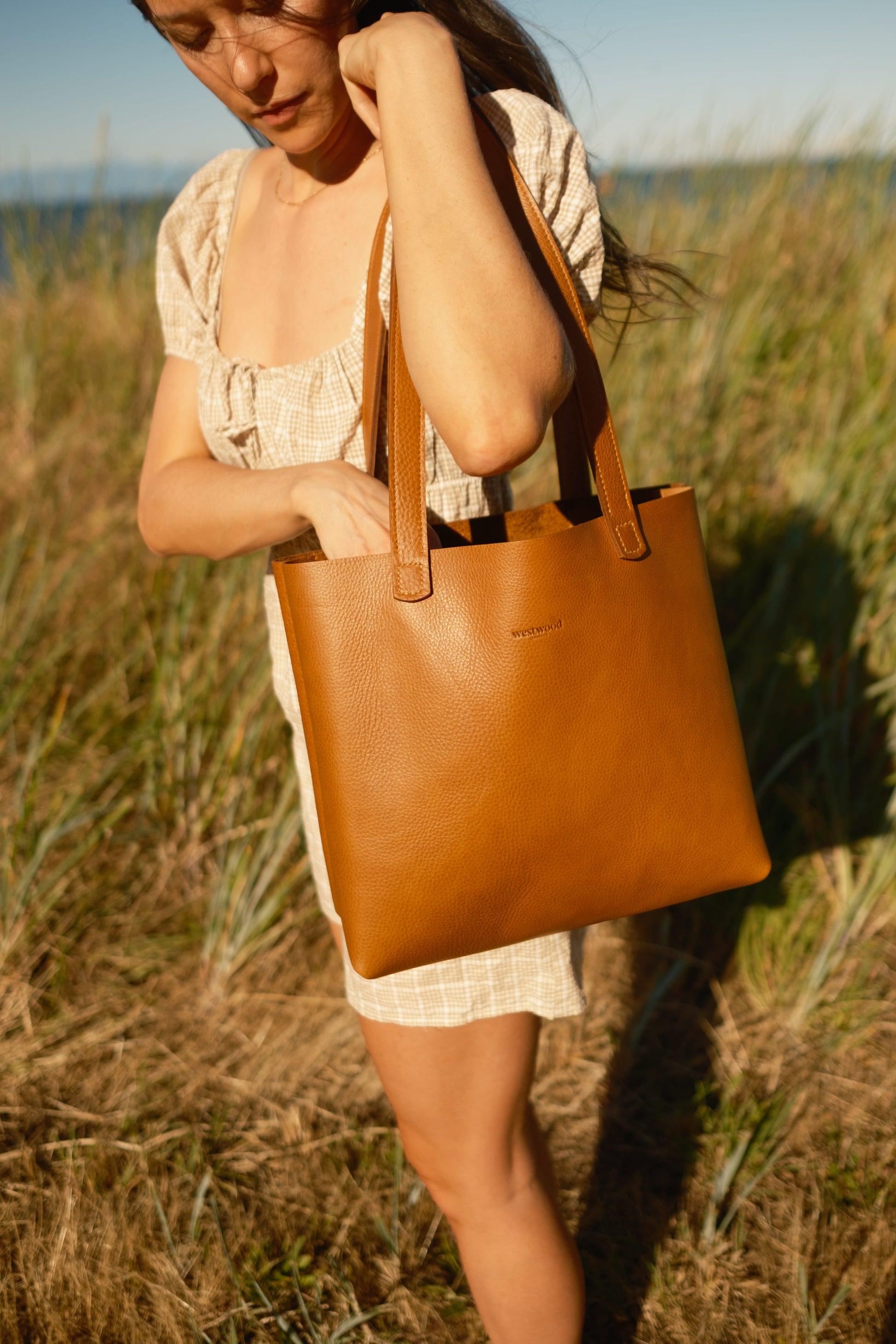 Woman holding a brown leather tote bag in a grassy field