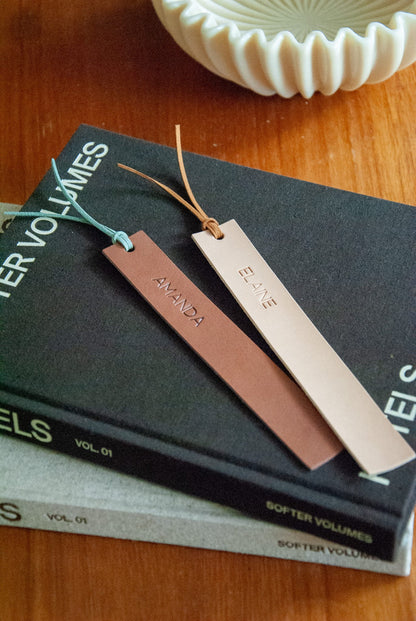 Two leather bookmarks on a stack of books with a decorative bowl in the background.