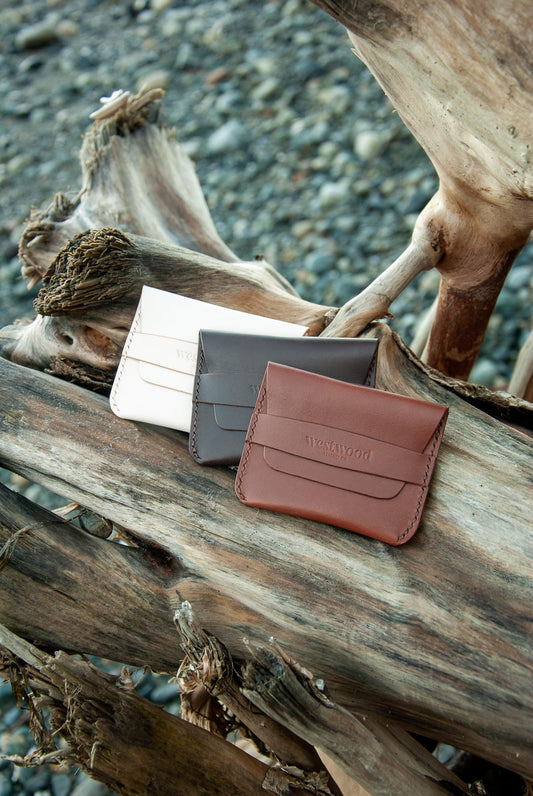 Three foldover flap wallets made of leather, placed on a wooden log. The wallets are in beige, black, and brown colors.