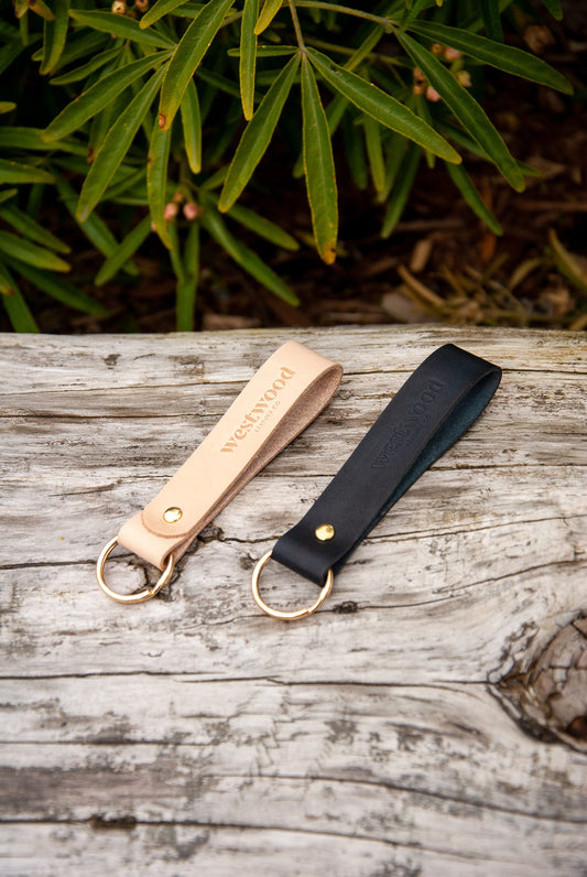 Two leather keychains, one beige and one black, with gold key rings, laid on a wooden surface with foliage in the background.