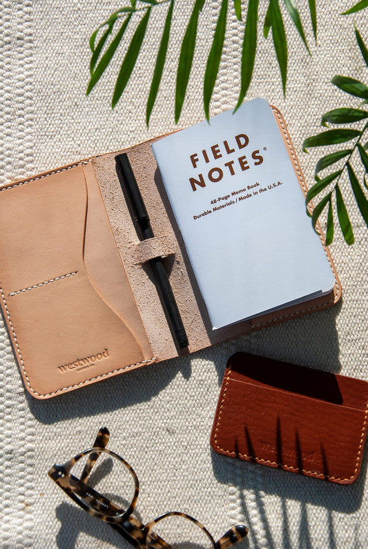 A brown leather journal cover displayed open with a Field Notes book inside, placed on a light-colored surface with a pair of glasses to the side.