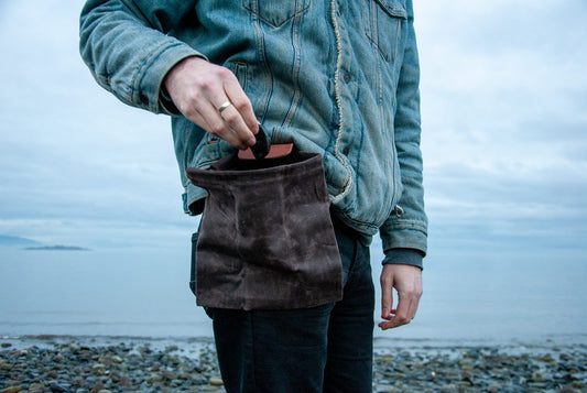 A person wearing a blue jacket and black pants is holding a brown foraging pouch with a leather strap and brass snaps.