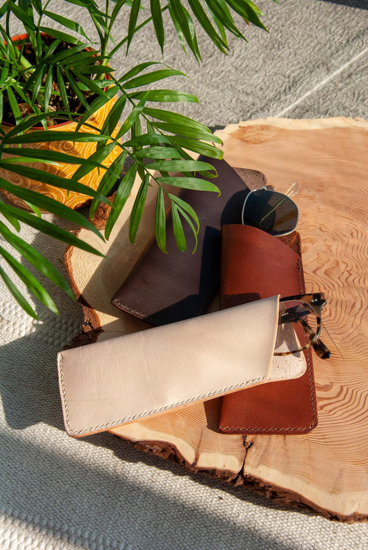 A brown glasses sleeve displayed on a wooden surface, with a pair of glasses partially inserted into it, alongside a monogram stamping tool and a plant pot in the background.