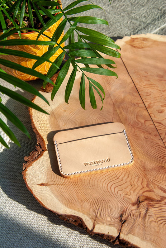 A slim beige card holder wallet made from vegetable tanned leather, displayed on a wooden surface with a green plant in the background.