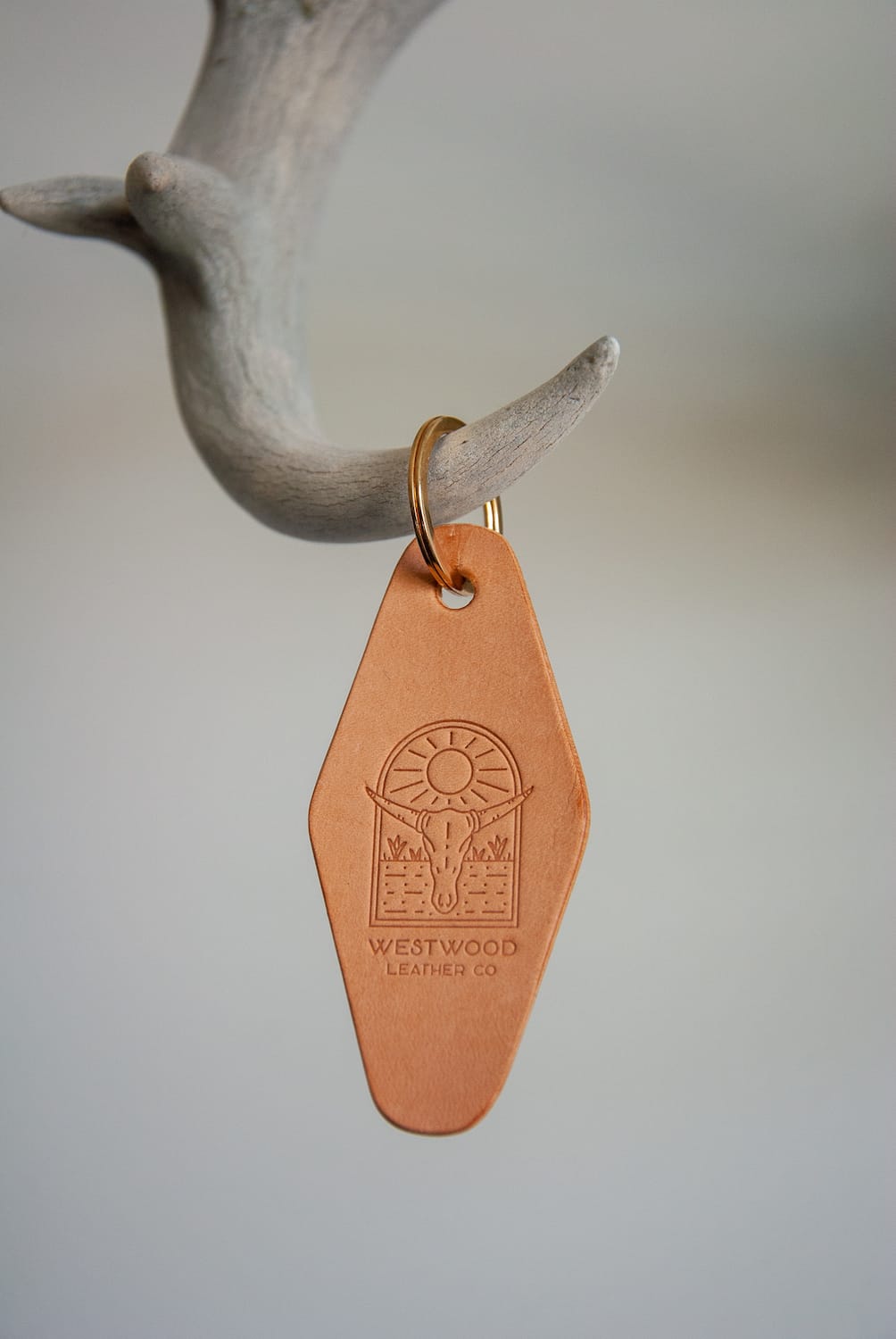A brown leather keychain with a brass key ring, hanging from a white branch. It features a stamped logo or design, possibly for a vintage motel theme.