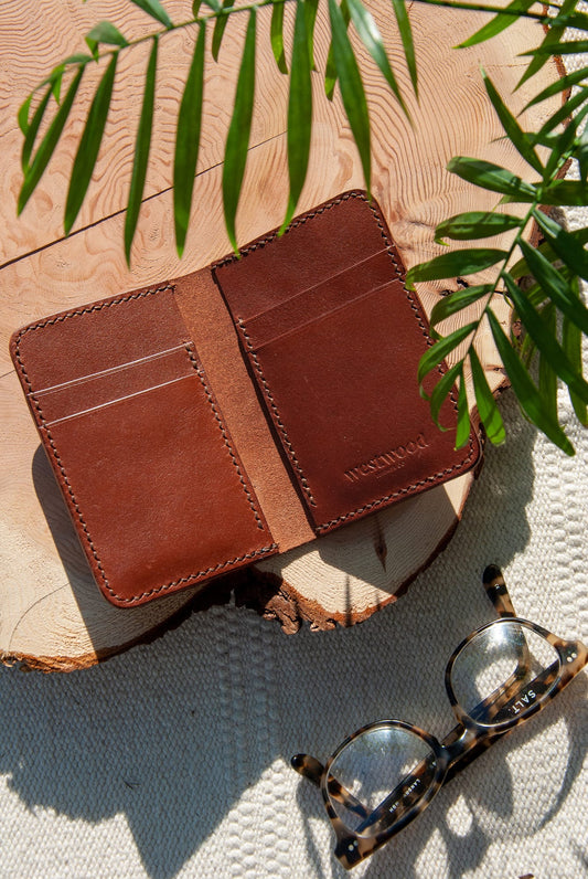 A brown bifold-style wallet with vertical card slots displayed open on a wooden surface, alongside a pair of glasses with a tortoiseshell pattern.